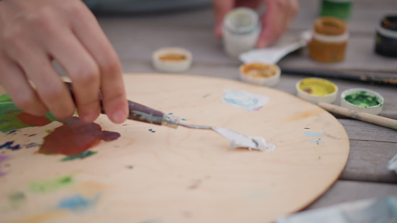 mani che mescolano pigmenti su una palette di legno, primo piano di dita che aprono piccoli barattoli di guazzo, spatola che stende pasta bianca, barattoli con toni della terra e verdi, giacca casual e jeans visibili