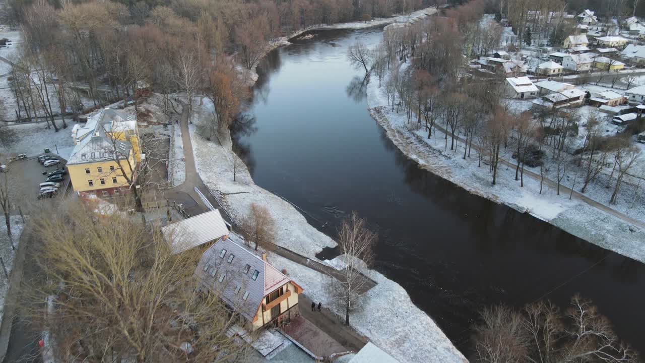 A breathtaking aerial view of Valmiera city, showcasing the winding Gauja River surrounded by snow-covered landscapes and residential areas under a wintry sky.