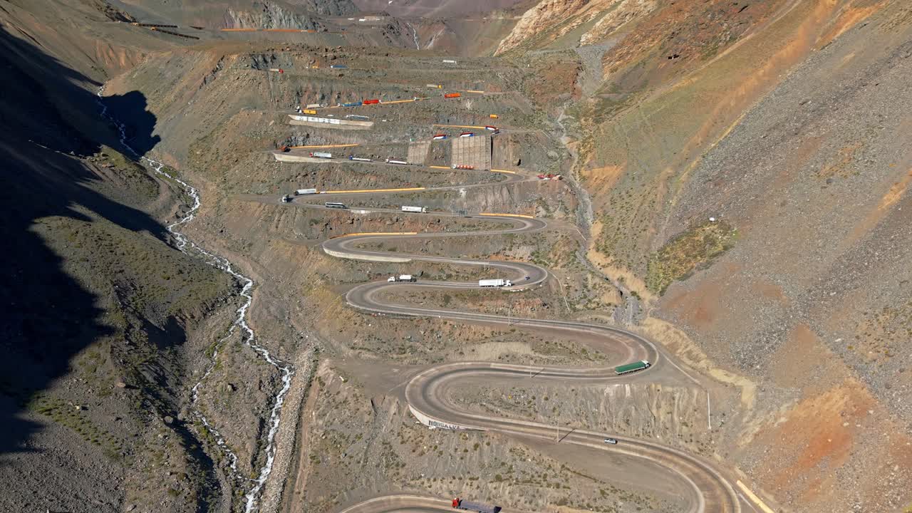 Aerial establishing view of Cuesta Caracoles hill with heavy trucks going up and down its curves on a sunny day