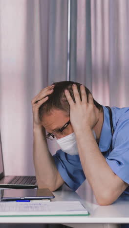 frustrated doctor in glasses wearing blue uniform and protective mask looks at tablet and holds head sitting at table with papers in hospital office