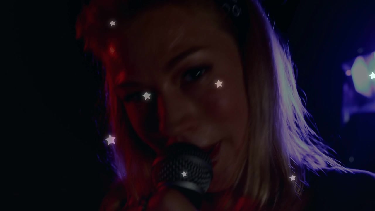 Cantante donna che entra nell'inquadratura e porta il microfono, cantando intensamente con un overlay a forma di stella che le attraversa il viso