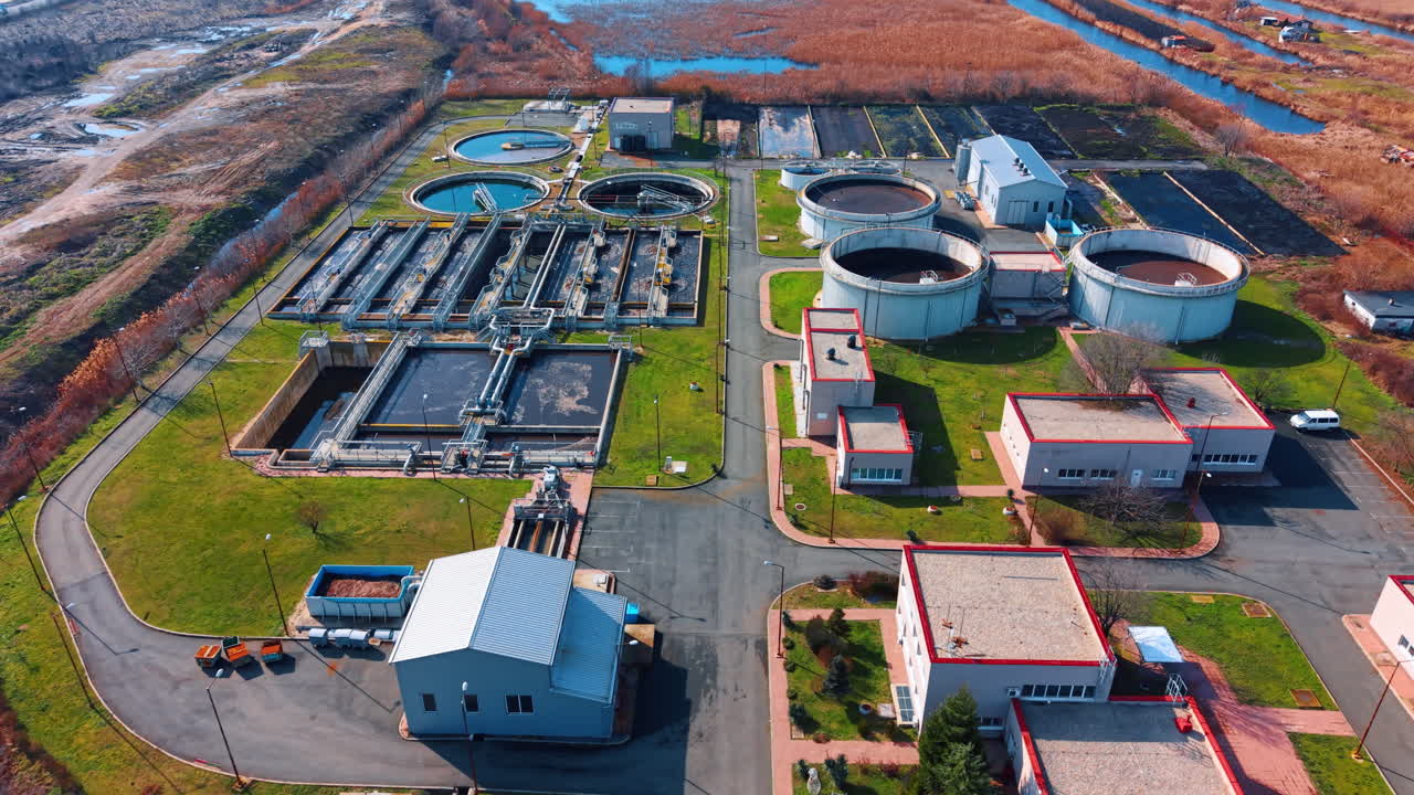 Wide aerial shot of modern sewage treatment plant infrastructure. Overview of circular clarifiers and rectangular sedimentation tanks near wetlands at daytime