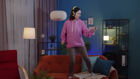 mujer adulta feliz con auriculares bailando y saltando mientras escucha música en casa sola en el sofá