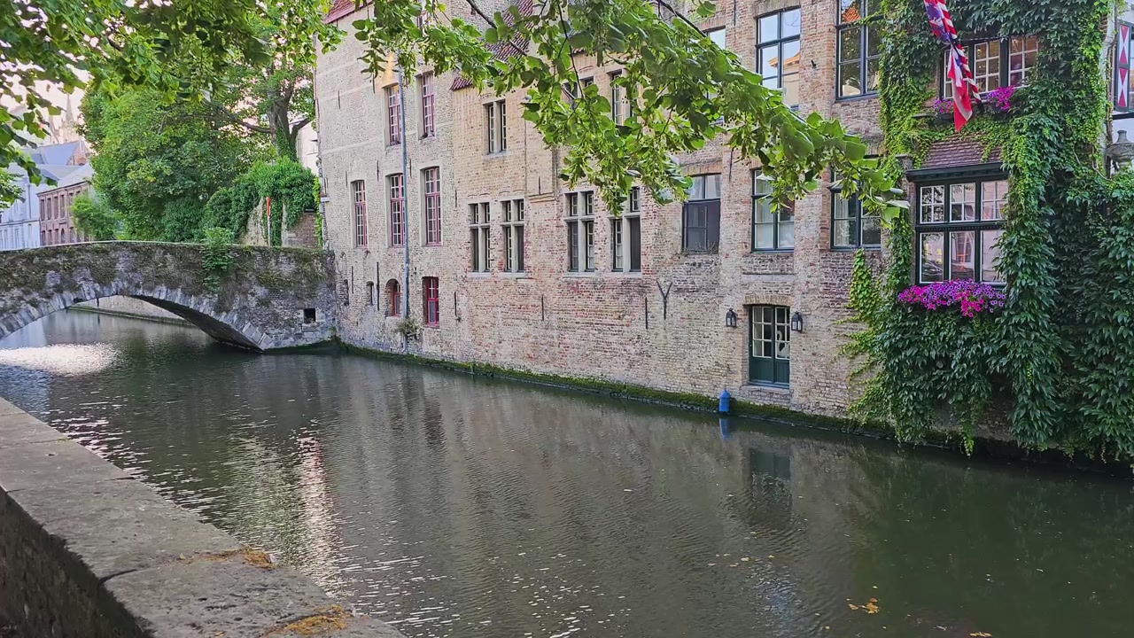 Charmerende udsigt over en gammel by ved floden med historiske murstenshuse, en buet stenbro, grønne slyngplanter, blomsterkasser, løvrige grene og stille kanalrefleksioner i et klassisk europæisk kanalmiljø.
