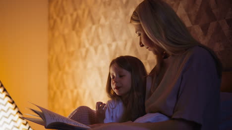 una niña feliz lee un libro sentada junto a su madre en la cama por la noche