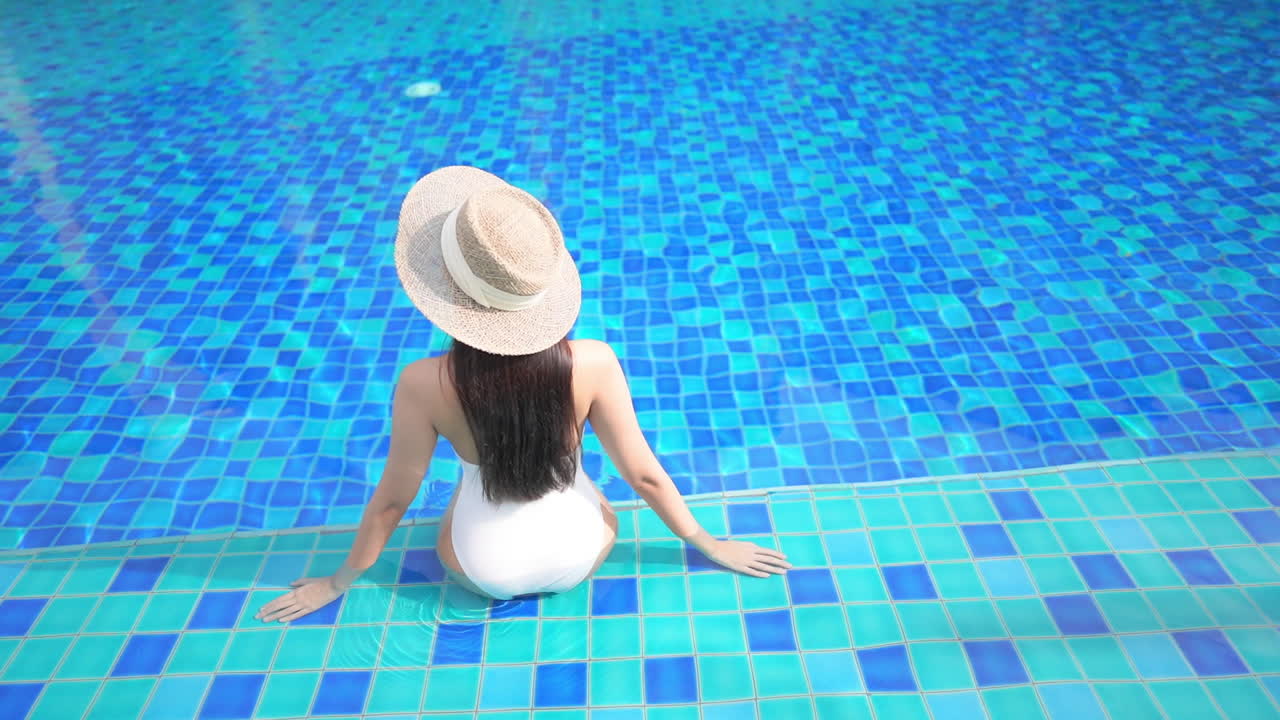 Back of Woman In Swimsuit and Summer Hat Sitting on a Pool Edge, High Angle View, slow-motion vacation template
