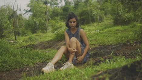 Tan-woman-sits-on-dirt-in-grassy-hillside-looking-down,-playing-with-dress