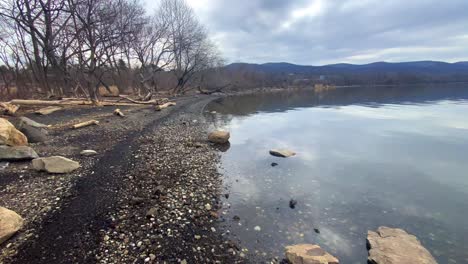 Panorámica-Sobre-La-Orilla-Rocosa-De-Un-Río-Con-árboles-Retorcidos,-Un-Vasto-Río-Y-Montañas-En-La-Distancia