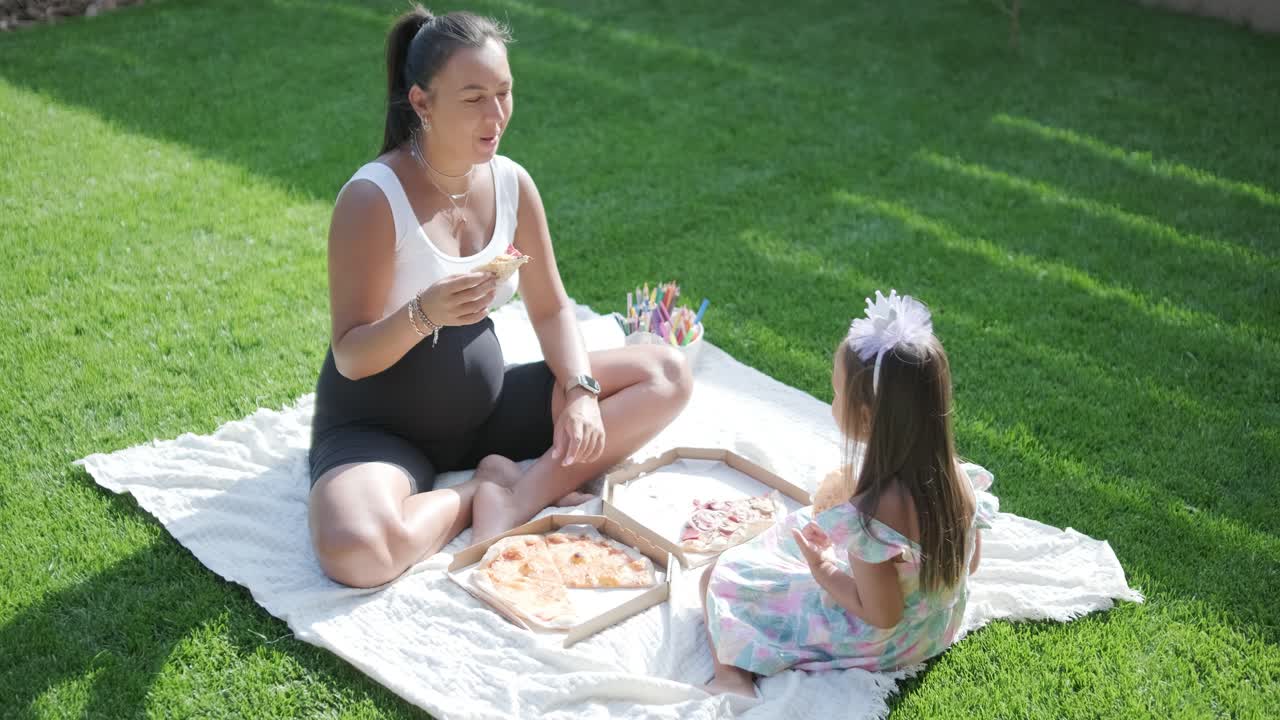 어머니와 딸은 잔디로 인 잔디에서 야외에서 여름 피크닉을 즐기고, 피자를 나누고, 은 날에 함께 좋은 시간을 보내고 있습니다. 가족 결합 장면은 여가와 휴식을 반영합니다.