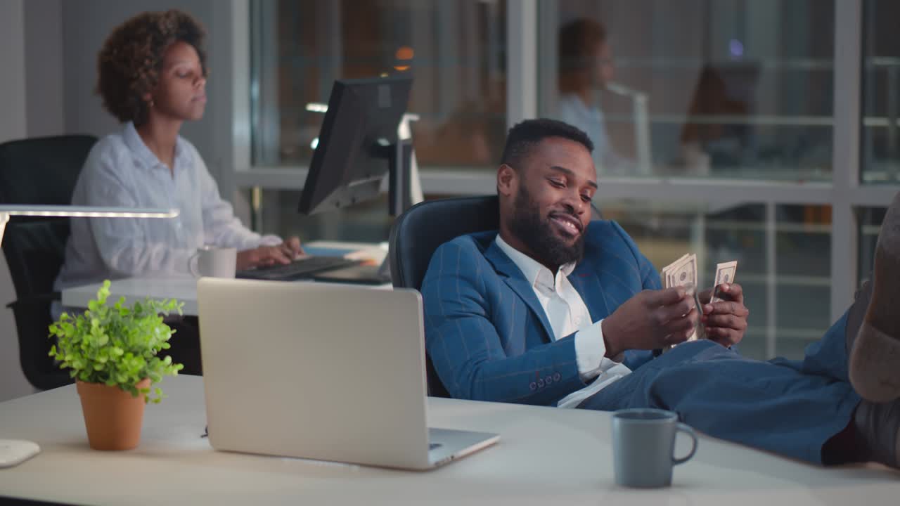 Young african businessman sitting at desk and counting money with colleague working on background