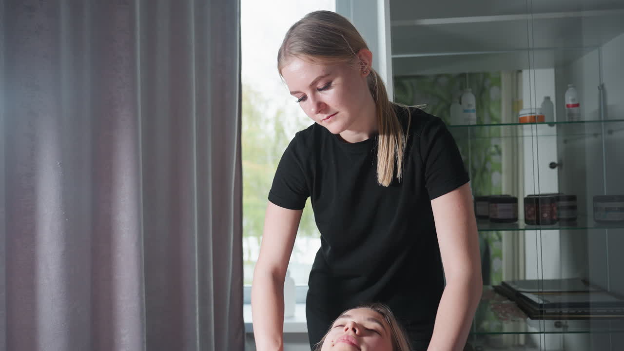 Massage therapist in black uniform providing chest massage to light skin client lying relaxed under white towel, with soft daylight entering beside grey curtain and skincare products on glass shelf