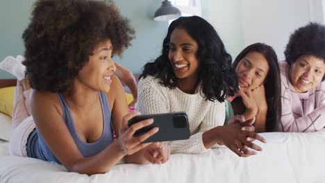 felices amigas diversas acostadas en la cama y usando el teléfono inteligente en el dormitorio