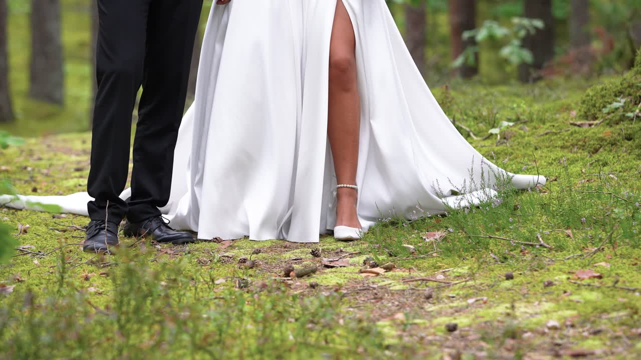 A couple gracefully walks along a mossy forest path, the bride's flowing white dress contrasting with the greenery. Soft natural lighting enhances this romantic and serene moment