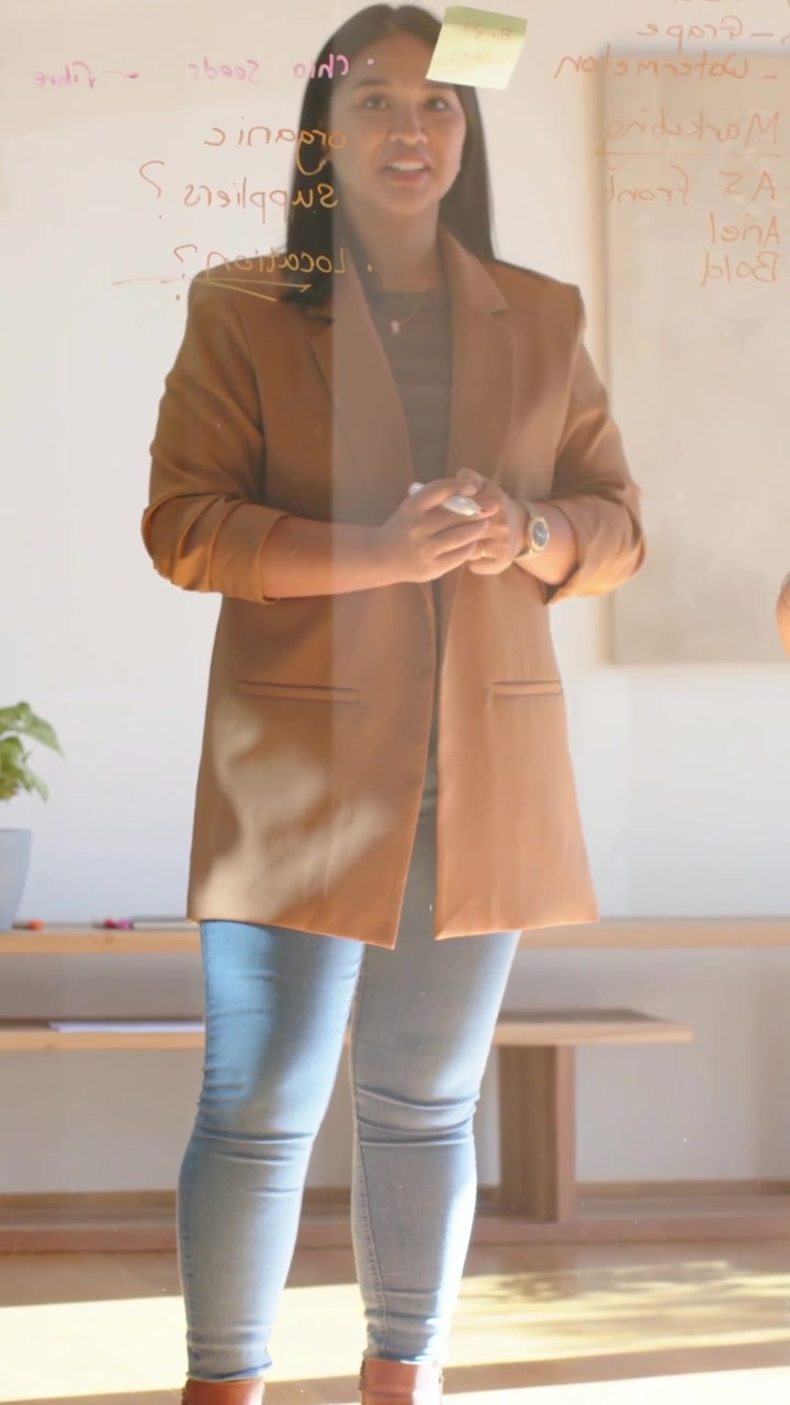 Video verticale: Donna in piedi con cappotto marrone che reagisce a un'indicazione su una lavagna di vetro con un post-it.