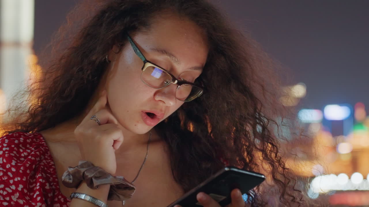 Biracial woman scrolling phone city lights, bokeh skyline background, red dress, glasses, reflective screen glow on face, thoughtful expression, tapping and typing, outdoor rooftop atmosphere, urban