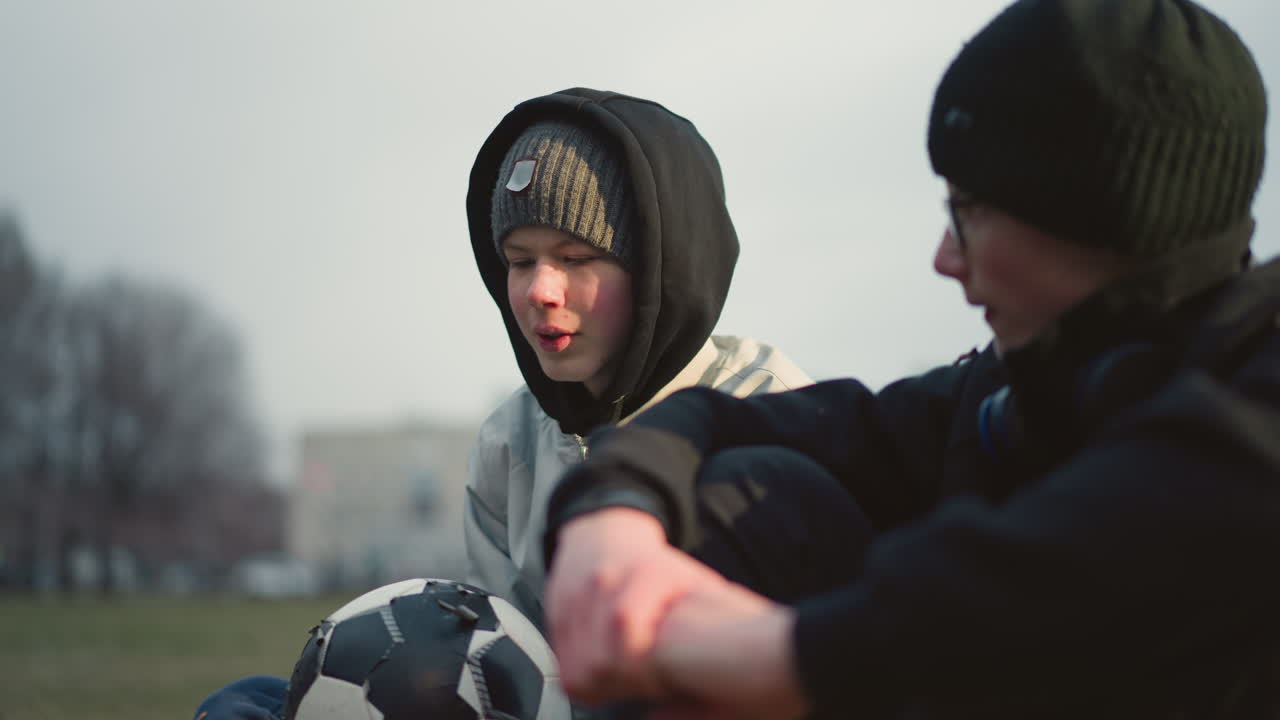 Close-up of two boys engaged in a discussion while sitting on a grassy field, the boy in a gray beanie and hoodie holds a soccer ball while the other in black clothing listens intently