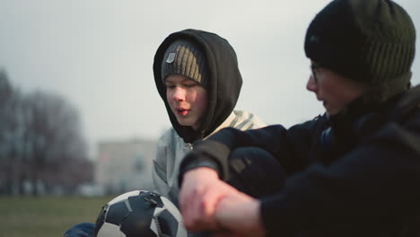 close-up of two boys engaged in a discussion while sitting on a grassy field, the boy in a gray beanie and hoodie holds a soccer ball while the other in black clothing listens intently