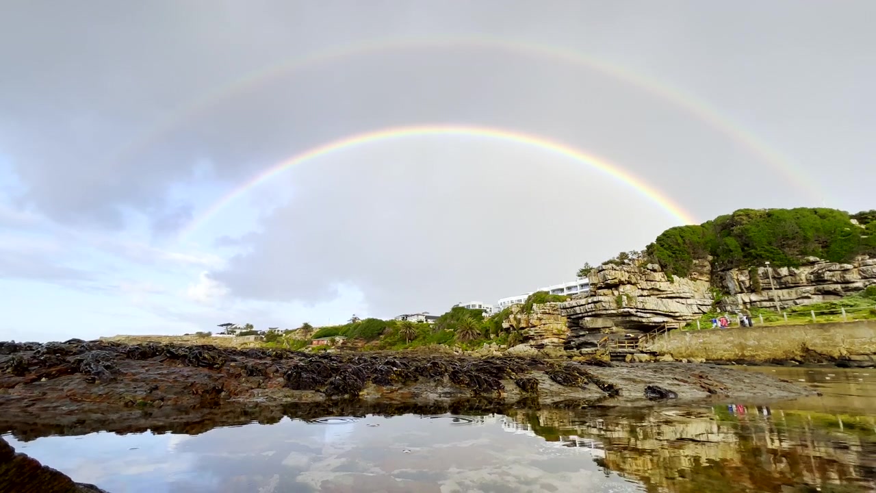 ハーマナスの潮だまりの穏やかな水面の上に、空にかかるカラフルな二重の虹