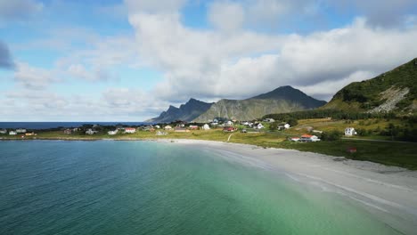 volando a lo largo de la playa paraíso en lofoten noruega con vista hacia los picos de las altas montañas con olas rompiendo a lo largo de las costas de la playa de arena blanca con casas rojas del norte a lo largo de la costa