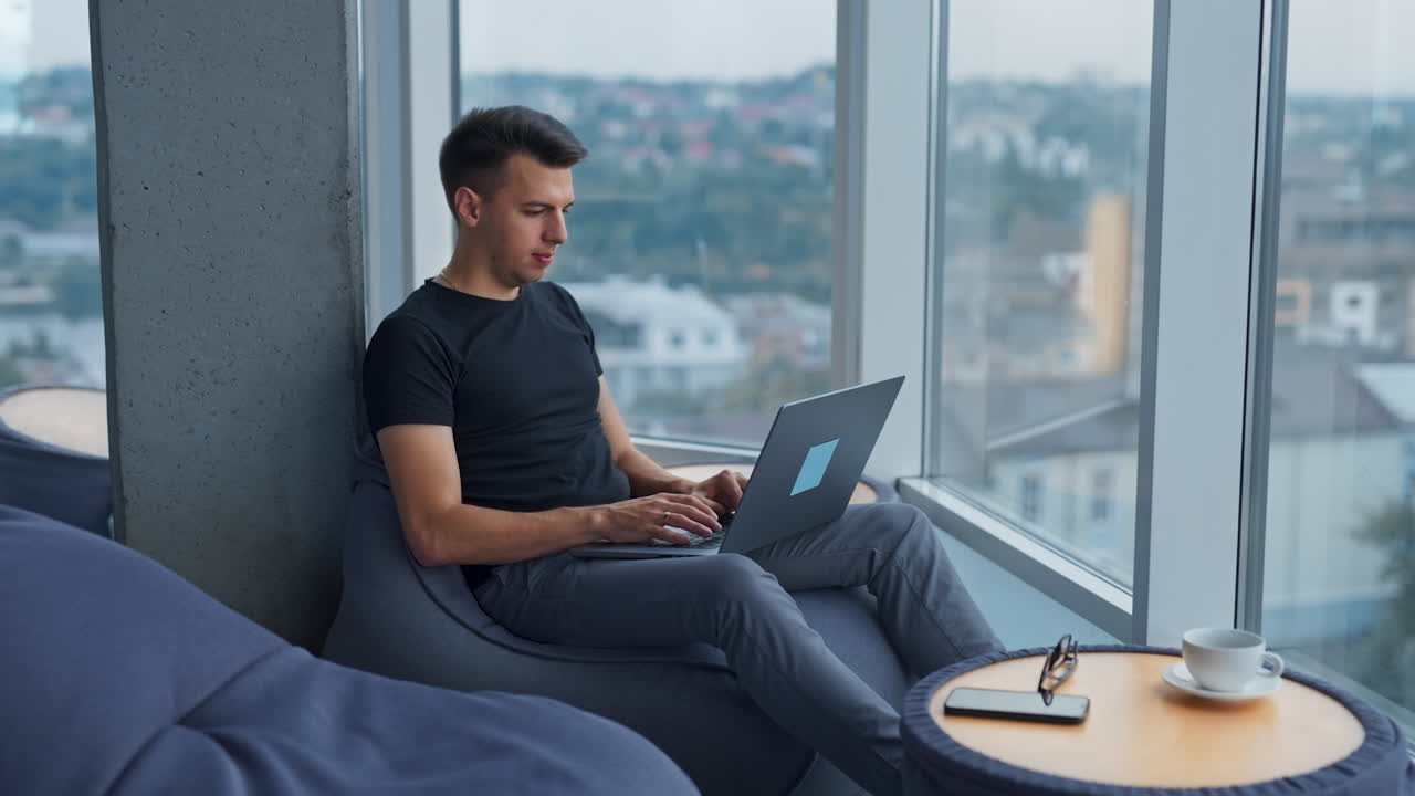 Young male freelancer working on laptop sitting comfortably near the panoramic window. Side view. Cityscape at backdrop in blur.
