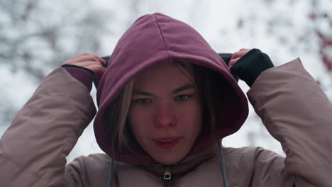 primer plano de una mujer fría ajustando la gorra de la capucha para mantenerse caliente en el frío clima invernal, el aire helado enrojece su cara mientras asegura su capucha, atmósfera invernal borrosa con ramas de árboles desnudas en el fondo