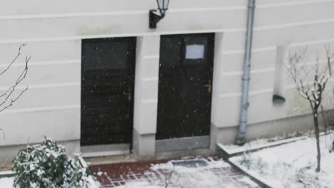 Snow-slowly-falling-on-ground-in-front-of-white-building-with-two-black-doors
