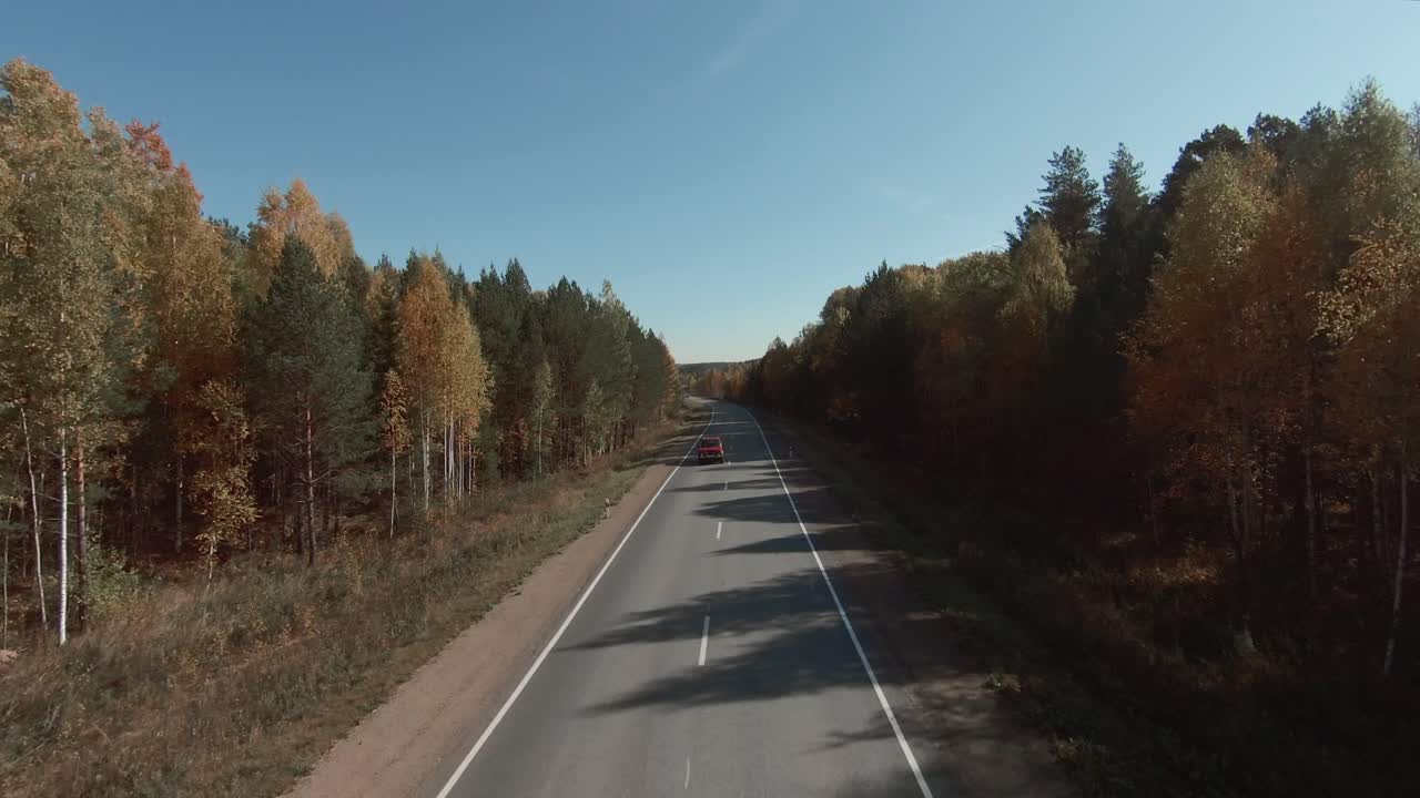 Riprese aeree FPV che sorvolano un'autostrada vuota a due corsie che attraversa una colorata foresta autunnale sotto un cielo azzurro e limpido, concetto di viaggio su strada rurale.