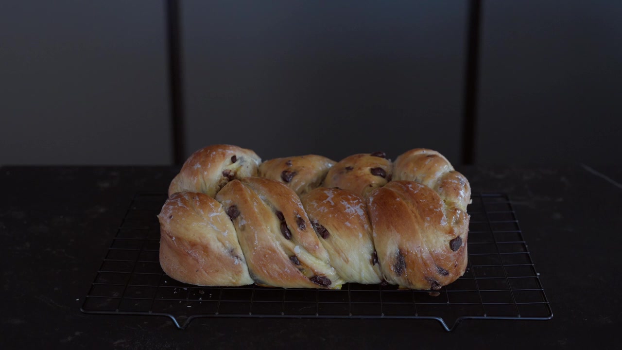 Slow tripod tilt down revealing a golden braided chocolate chip brioche resting on a wire rack over a dark countertop.