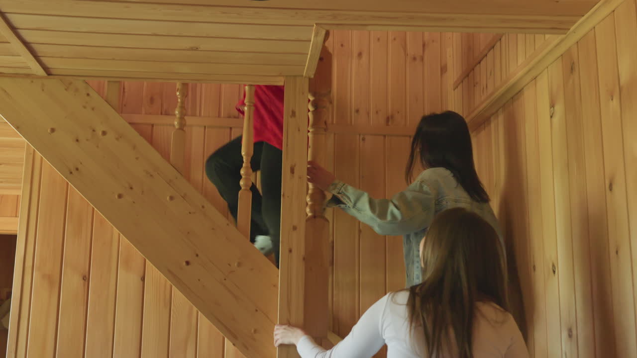group of cheerful women walking through wooden cabin living room as one girl in red top with sunglasses around neck leads friends toward staircase, smiling and talking in cozy relaxed home atmosphere
