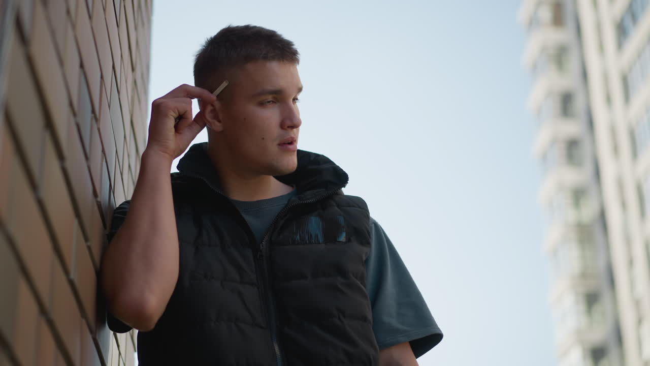 young smoker in dark vest stands against brick wall removing cigarette from ear preparing to light it with blurred view of modern tall building in background under clear sky