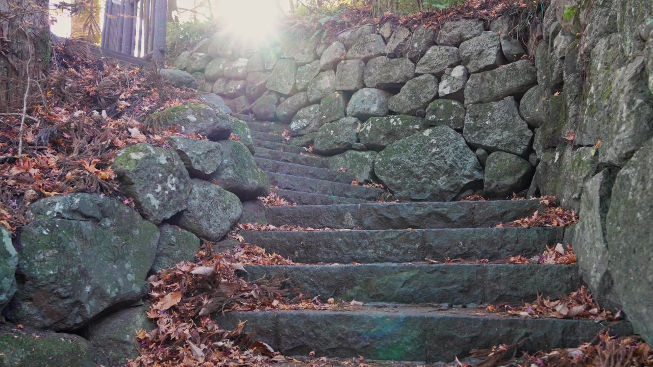 Ancient stone steps lead upward between weathered rock walls as warm sunlight creates a natural lens flare over fallen autumn leaves, evoking history, heritage and exploration.