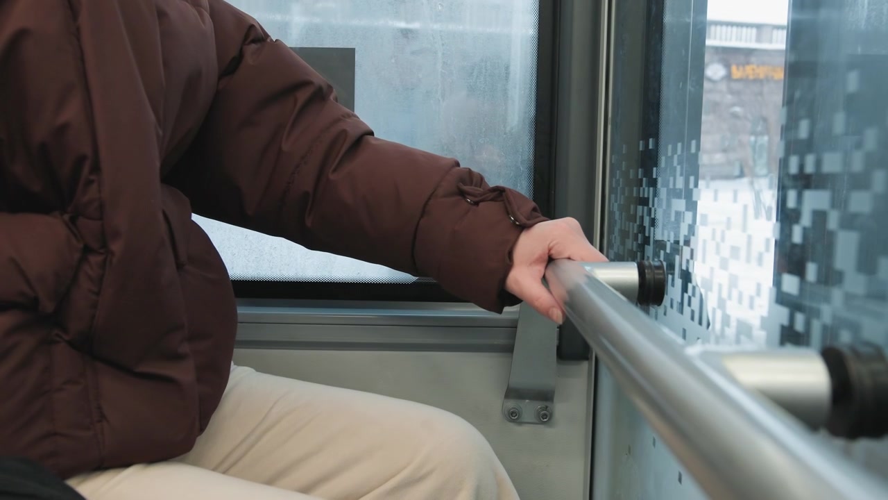 Vista cercana de la mano derecha con una chaqueta acolchada marrón sujetando una barra de agarre metálica junto a la condensación en la ventana dentro de un autobús o tren, transmitiendo equilibrio estable, comodidad invernal y el viaje urbano diario.