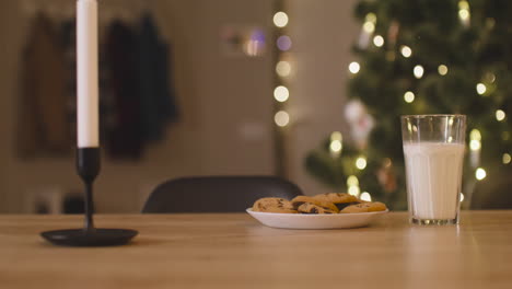 la cámara se enfoca en un vaso de leche y un plato lleno de galletas en una mesa vacía con dos velas en una habitación decorada con un árbol de navidad