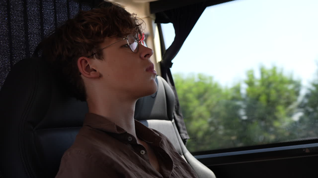 Young man with glasses sitting in a vehicle, gazing thoughtfully out the window during a journey