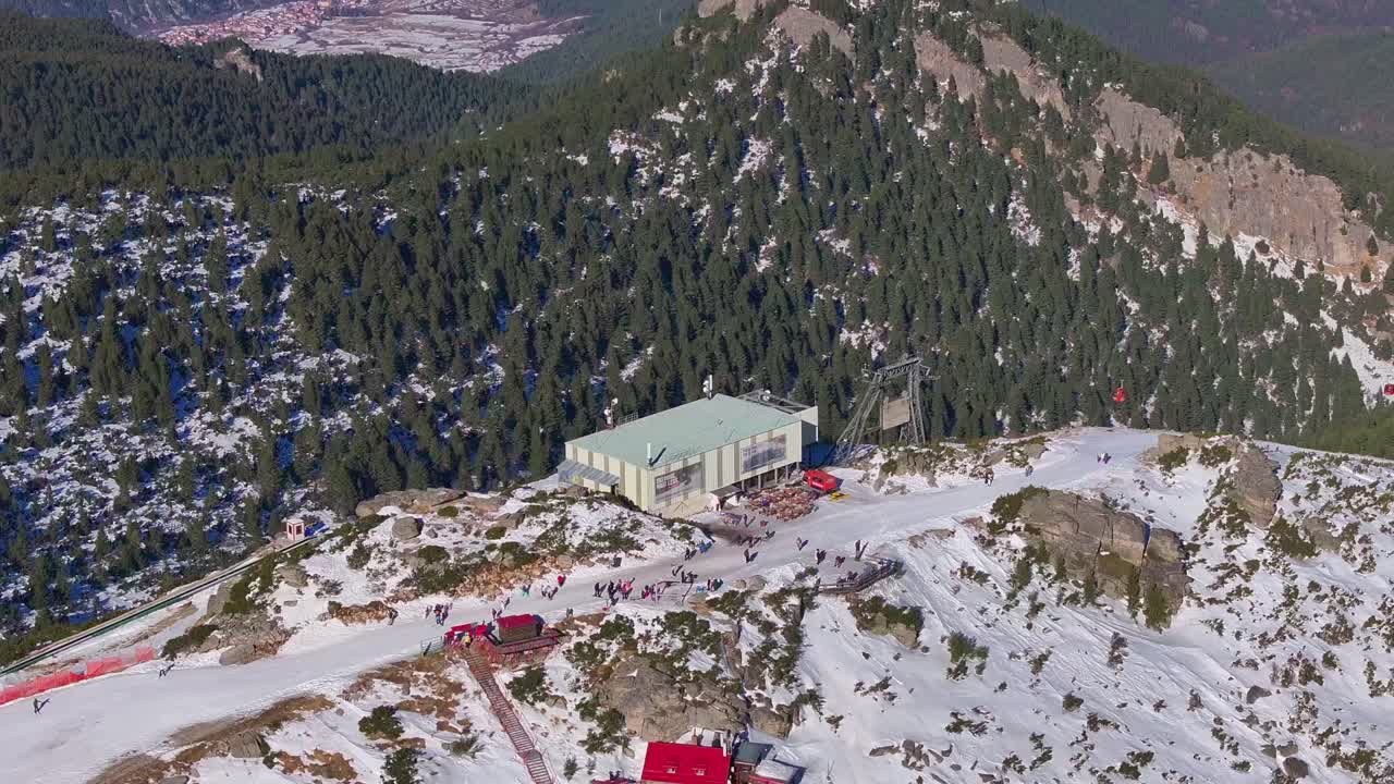 This video represent ski resort on the top of the mountain, lot of people skiing. Ski crowd. People using chairlifts. There is a lot of trees on the other mountain in the distance behind ski restort.