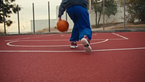 Close-up-shot-of-a-girl-in-blue-pants-hitting-an-orange-basketball-off-the-red-floor-on-a-basketball-court-and-running-to-throw-the-ball-into-the-basketball-hoop