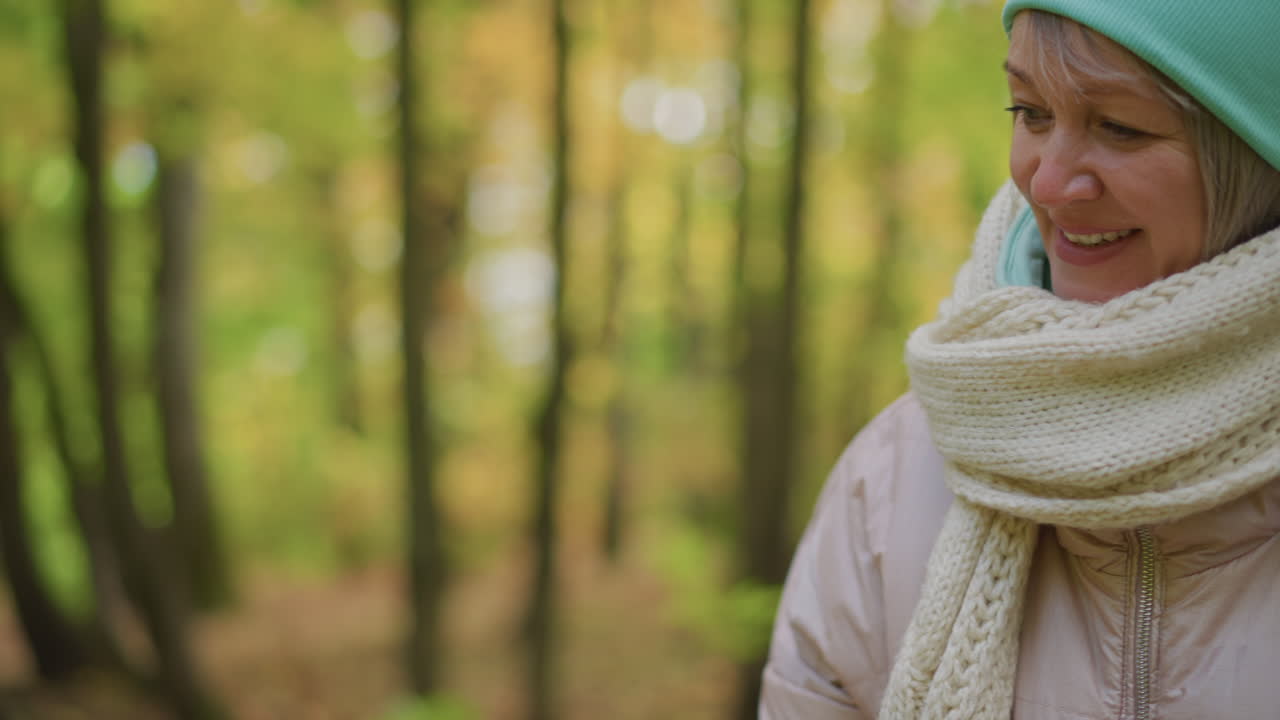 mother smiles warmly while handing bright autumn leaf to daughter in serene park, both dressed in cozy hats and jackets surrounded by soft light and blurred golden green fall forest background