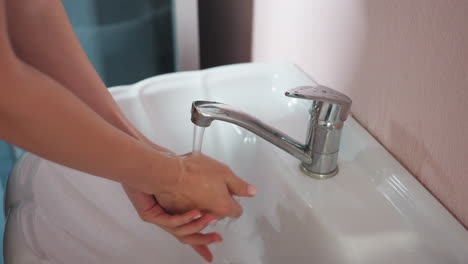 close-up of light skin woman washing hands under running water in white ceramic sink with silver tap, against peach-colored wall, with water passing cleanly through visible neat pan connector