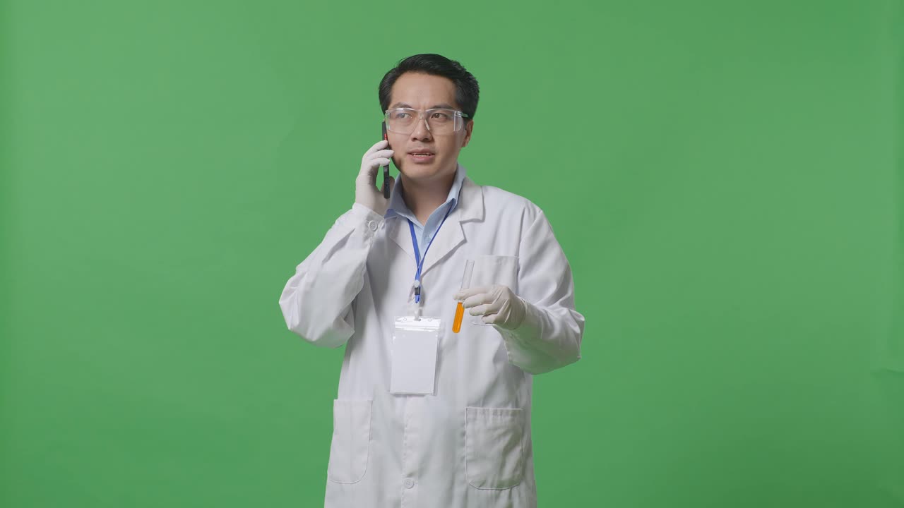 Asian Man Scientist Looking At The Orange Liquid In The Test Tube In His Hand And Talking On Smartphone While Standing On The Green Screen Background In The Laboratory