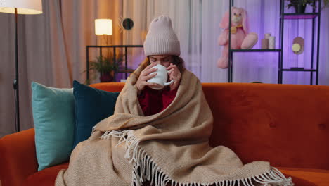 sick ill teen girl wear hat wrapped in plaid sitting, shivering from cold on sofa drinking hot cacao