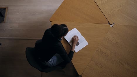 Imágenes-De-Vista-Superior-De-Una-Joven-Africana-Concentrada-Sentada-Junto-Al-Escritorio-Redondo-De-Madera-Y-Leyendo-Un-Libro,-Estudiando-Para-La-Universidad-En-La-Biblioteca-Moderna,-Preparándose-Para-Los-Exámenes.-Vistiendo-Un-Suéter-Negro