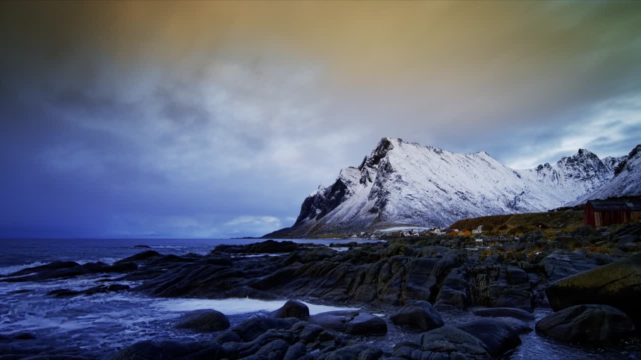 Floating clouds reflecting sunset moving from the background towards the foreground above snow-covered mountain behind the village by the rocky shore