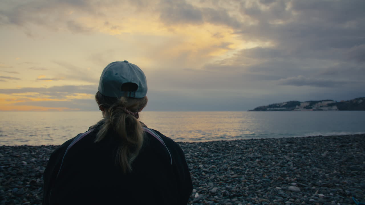 A woman sits facing the ocean during a dramatic sunset. Soft light, solitude, and quiet reflection on a rocky shoreline