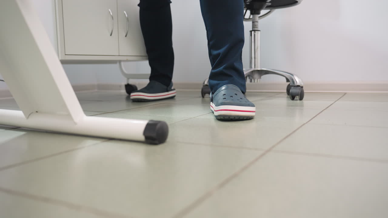 Leg view of treatment provider wearing crocs and navy blue uniform standing beside clinical bed under bright lamp with mobile stool, product table and cabinet visible on clean tiled floor