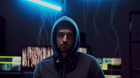 portrait of the young good looking man hacker in a hood looking straight to the camera while sitting in the dark server room with computers