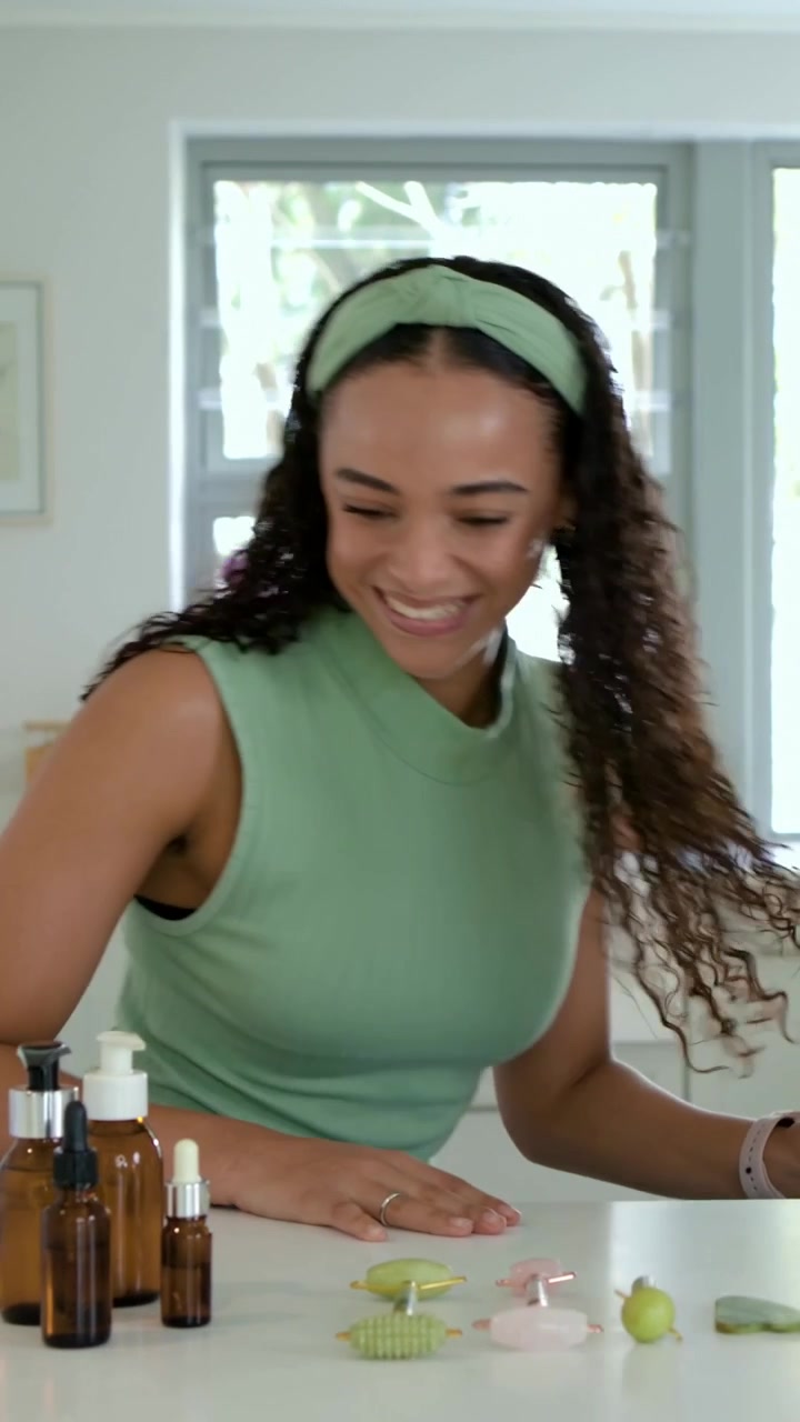 Video verticale: Donna con top verde che si allunga, allineando bottiglie ambrate e una maschera LED in cucina per una dimostrazione.