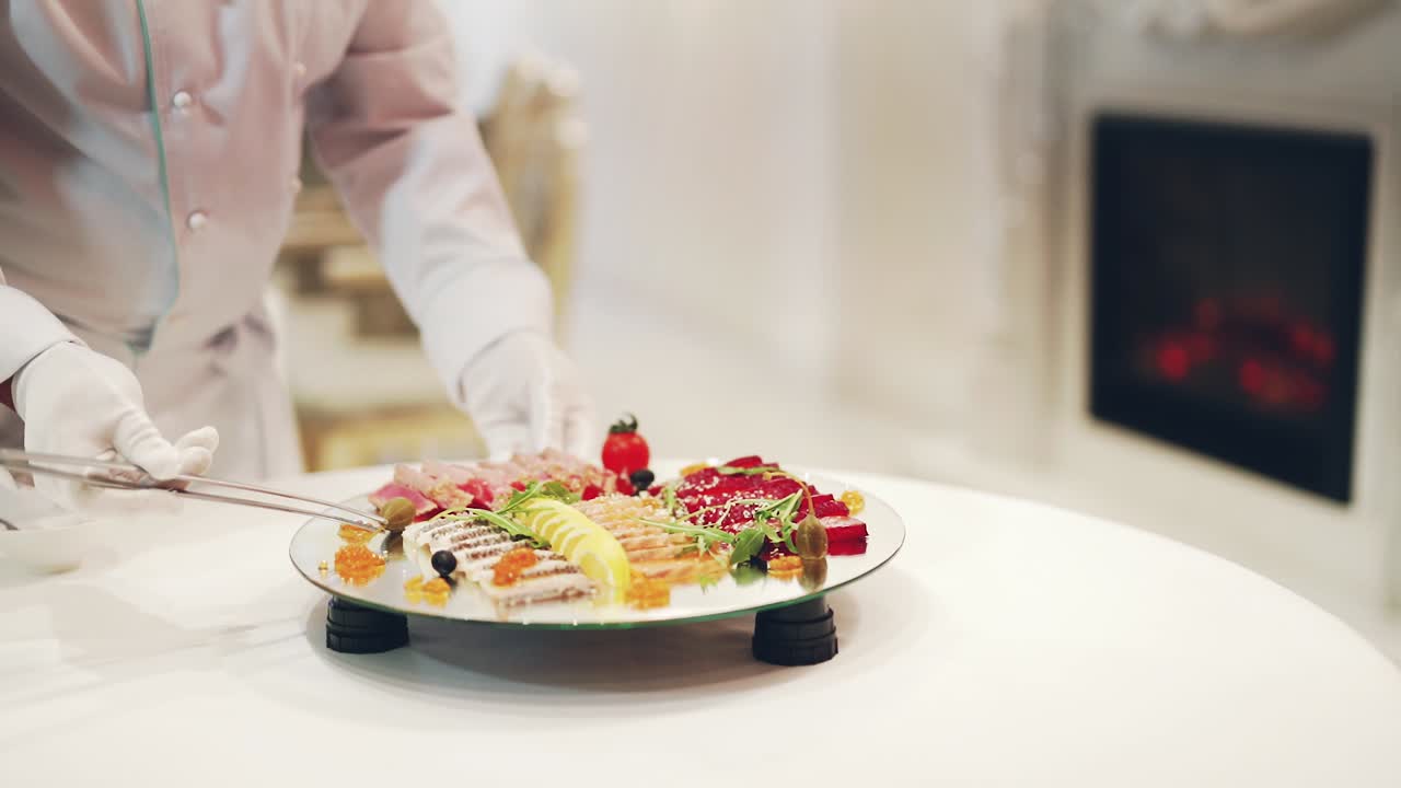 Unrecognizable male cook in white gloves decorates plate with delicious food on a glass turntable. Professional cook puts slicing products prettily for guests.