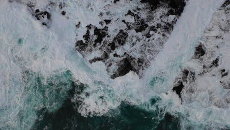 Aerial-zoom-out-as-ocean-waves-crash-against-a-rugged-coastline