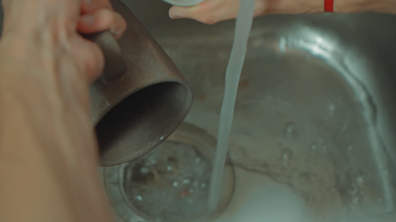 Close-up of a person's hands washing and rinsing two ceramic mugs under running tap water in a stainless steel kitchen sink