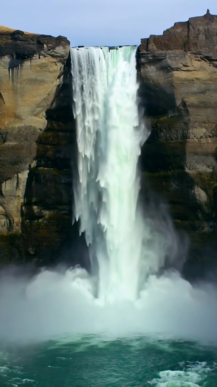 Video verticale: Acqua che si riversa su un bordo roccioso creando una cascata dalla scogliera in schiuma e nebbia, con una pozza d'acqua alla base.